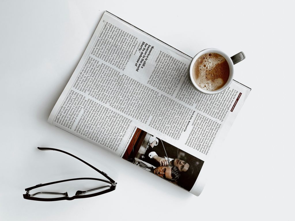 Blick von oben auf eine Zeitschrift, auf der ein Kaffee steht und neben der eine schwarze Brille liegt.